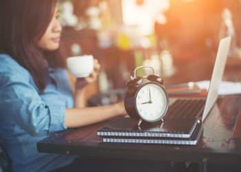 Mulher trabalhando com laptop e relógio analógico em primeiro plano — futuro dos trabalhadores de aplicativos e discussão sobre o fim da escala 6x1