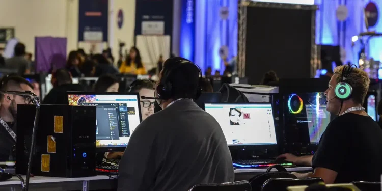 Grupo de profissionais com fones de ouvido trabalhando em computadores desktop durante evento de tecnologia, com iluminação azul ao fundo e diversas estações de trabalho ao longo do ambiente