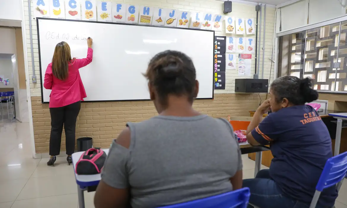 Estudantes adultos em sala de aula da EJA assistindo à professora escrever no quadro, representando o público que pode receber a Bolsa Presença de R$ 100 por frequência escolar