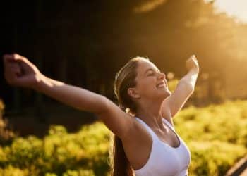 Mulher sorrindo e comemorando sua conquista ao ar livre, com os braços levantados em sinal de vitória.