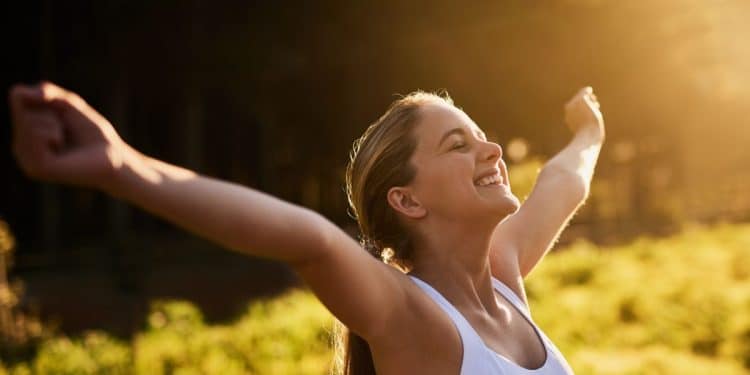 Mulher sorrindo e comemorando sua conquista ao ar livre, com os braços levantados em sinal de vitória.