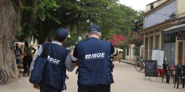Agentes do IBGE de costas caminhando por rua com coletes azuis identificados com o logotipo do Instituto Brasileiro de Geografia e Estatística