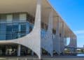 Vista externa lateral do Palácio do Planalto em Brasília, destacando suas colunas brancas icônicas, fachada de vidro e o céu azul ao fundo.