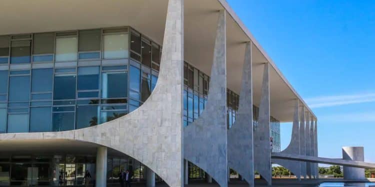 Vista externa lateral do Palácio do Planalto em Brasília, destacando suas colunas brancas icônicas, fachada de vidro e o céu azul ao fundo.