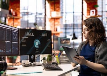 Mulher analisando gráficos em monitores e utilizando tablet em escritório moderno