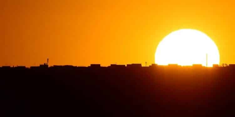 Fotografia de paisagem urbana ao pôr do sol ou nascer do sol. O céu está predominantemente em tons intensos de laranja, amarelo e ouro. À direita, um sol circular muito grande, branco e brilhante, começa a se esconder atrás da linha do horizonte. O horizonte é uma silhueta preta e sólida de edifícios, prédios baixos e antenas de comunicação, sem detalhes visíveis. A parte inferior da imagem é composta por uma área escura, preta e indefinida.