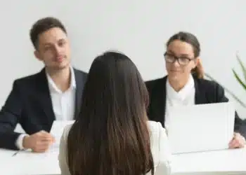 Candidata de costas sendo entrevistada por dois recrutadores em ambiente corporativo, representando o momento da entrevista de emprego para o qual a Inteligência Artificial pode ajudar na preparação