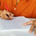 Pessoa escrevendo com caneta vermelha em caderno quadriculado representando o preenchimento da inscrição para o concurso da UFMA com 59 vagas.