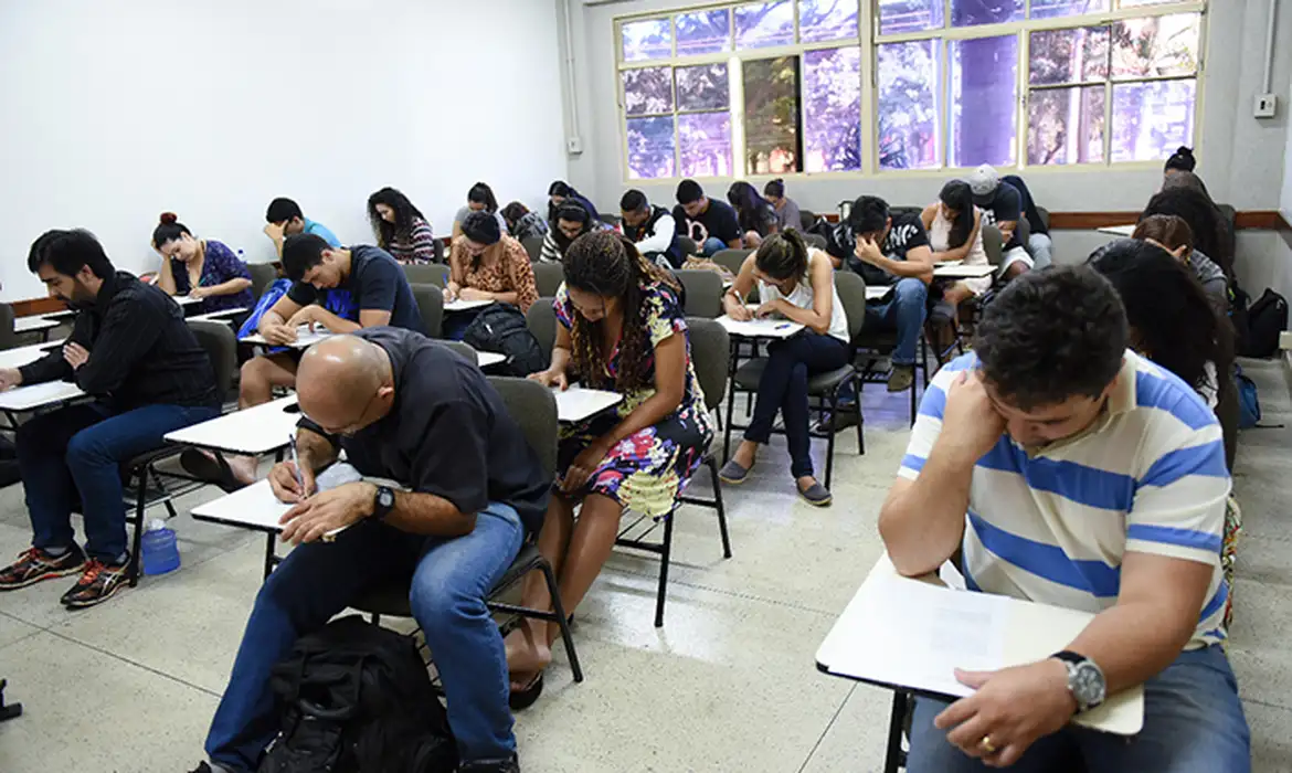 Sala de aula com adultos e jovens sentados em carteiras universitárias realizando prova escrita, com janelas amplas ao fundo deixando entrar luz natural, representando a aplicação do Encceja para certificação do ensino fundamental e médio
