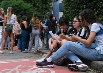Grupo de jovens estudantes reunidos do lado de fora de instituição de ensino, com três deles sentados na calçada analisando um documento, em ambiente com árvores e movimento de pessoas ao fundo, ilustrando estudantes universitários em processo de inscrição no programa Pé-de-Meia Licenciaturas 2026