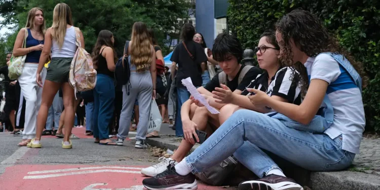 Grupo de jovens estudantes reunidos do lado de fora de instituição de ensino, com três deles sentados na calçada analisando um documento, em ambiente com árvores e movimento de pessoas ao fundo, ilustrando estudantes universitários em processo de inscrição no programa Pé-de-Meia Licenciaturas 2026