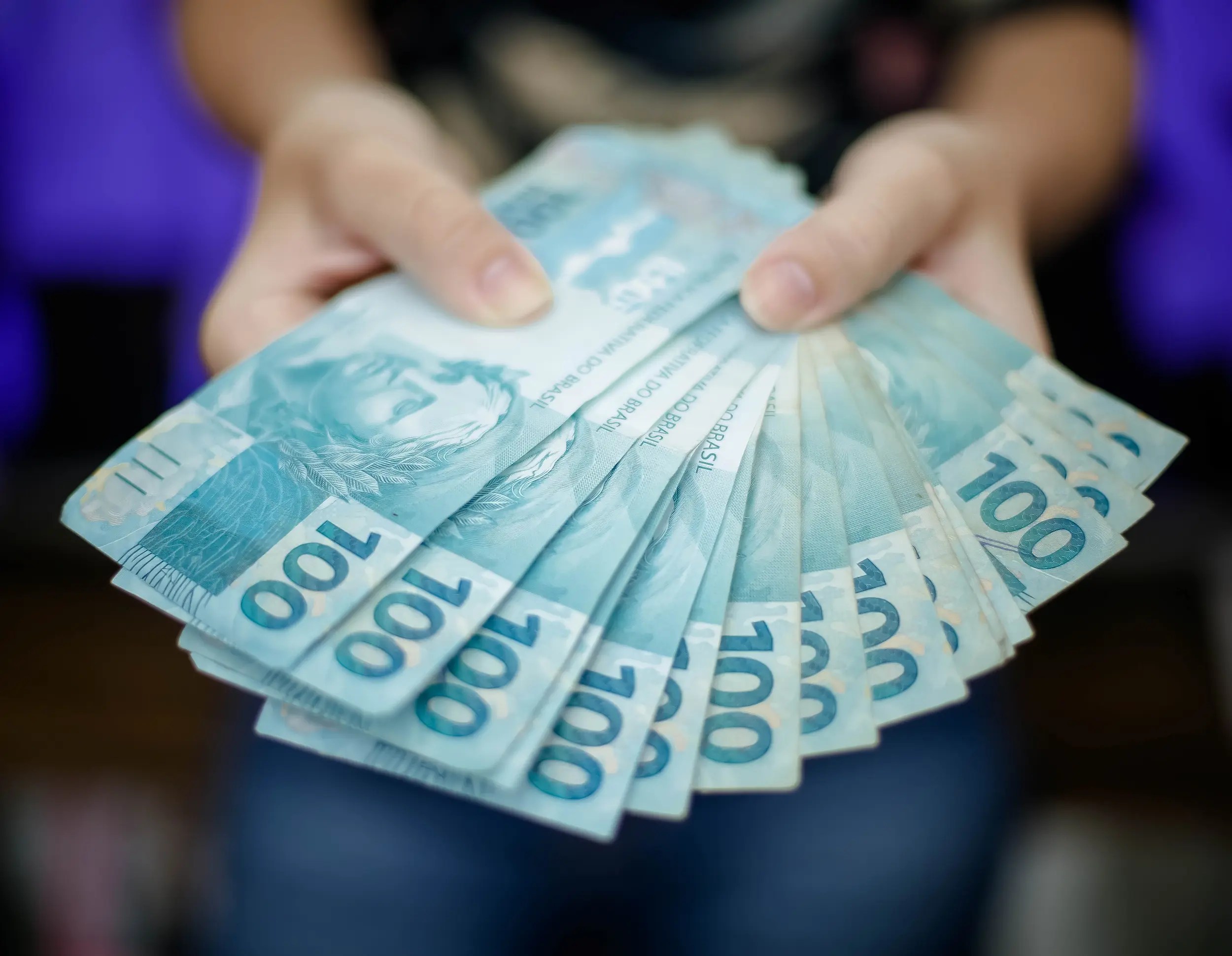 Notas de R$ 100,00 em mãos, representando o pagamento de R$ 8.475,55.