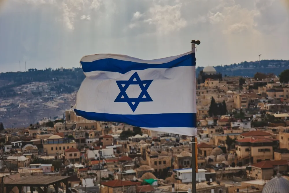 Bandeira de Israel tremulando sobre cidade histórica com construções urbanas ao fundo representando o povo israelense