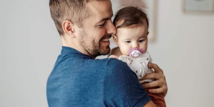 Um homem de camiseta azul sorri enquanto segura um bebê no colo. O bebê está usando uma chupeta e olha atentamente para o lado em um ambiente doméstico acolhedor.