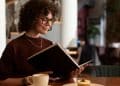 Mulher com óculos e cabelo cacheado lendo um livro em um café, representando o hábito da leitura como ferramenta para desenvolver a inteligência emocional e o bem-estar mental.