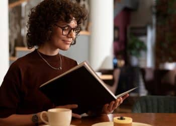 Mulher com óculos e cabelo cacheado lendo um livro em um café, representando o hábito da leitura como ferramenta para desenvolver a inteligência emocional e o bem-estar mental.
