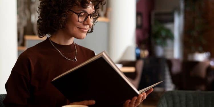 Mulher com óculos e cabelo cacheado lendo um livro em um café, representando o hábito da leitura como ferramenta para desenvolver a inteligência emocional e o bem-estar mental.