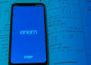 Smartphone exibindo tela do ENEM apoiado sobre livro aberto de matemática com questões