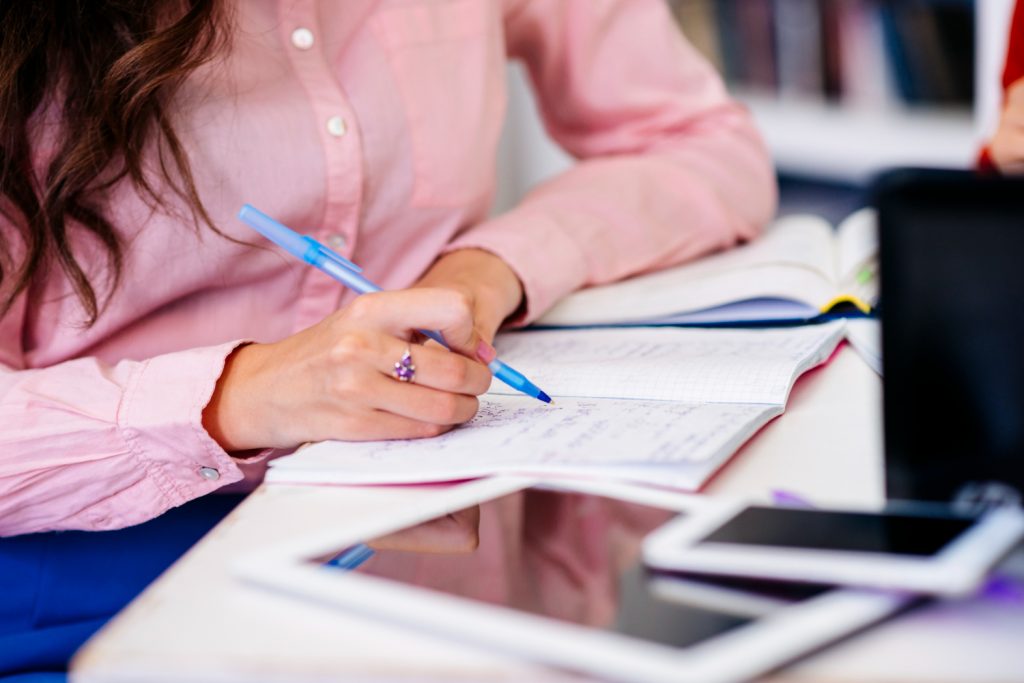 Mão feminina com caneta azul anotando em caderno aberto sobre mesa com tablet e smartphone ao lado em sessão de estudos