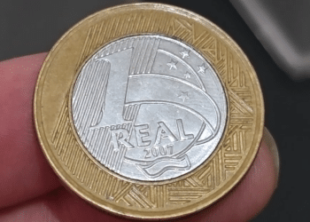 Uma pessoa segurando uma moeda de 1 Real de 2007 entre o polegar e o indicador, mostrando o detalhe do valor e do ano em um close fotográfico.