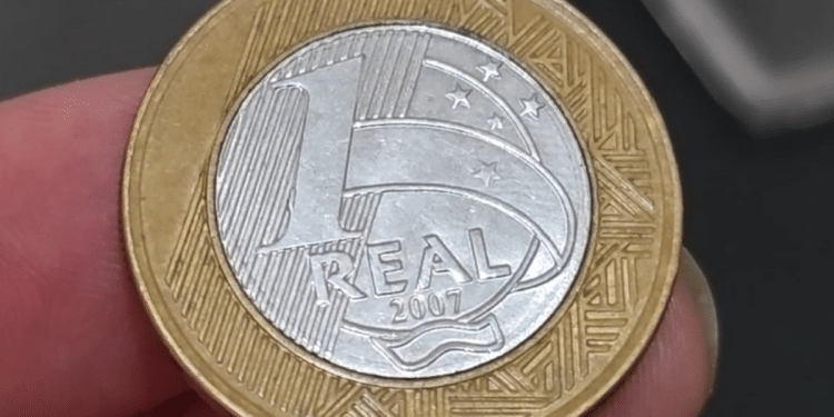 Uma pessoa segurando uma moeda de 1 Real de 2007 entre o polegar e o indicador, mostrando o detalhe do valor e do ano em um close fotográfico.