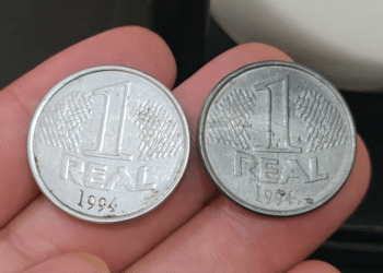 Duas moedas de 1 real de 1994 seguradas entre os dedos; a da esquerda é brilhante e prateada (original), enquanto a da direita possui um tom cinza mais escuro e fosco (variante falsa de época).