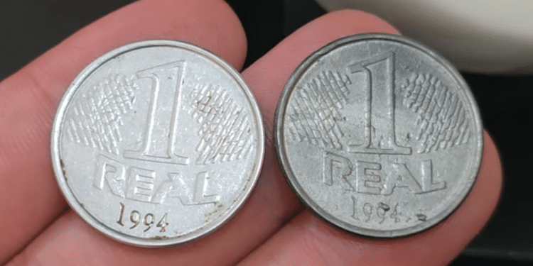 Duas moedas de 1 real de 1994 seguradas entre os dedos; a da esquerda é brilhante e prateada (original), enquanto a da direita possui um tom cinza mais escuro e fosco (variante falsa de época).