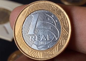 Close de uma mão segurando a moeda de 1 real comemorativa dos 50 anos do Banco Central, lançada em 2015, mostrando o lado do valor (reverso) com detalhes em dourado e prata.