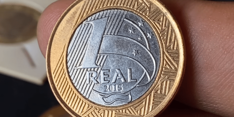 Close de uma mão segurando a moeda de 1 real comemorativa dos 50 anos do Banco Central, lançada em 2015, mostrando o lado do valor (reverso) com detalhes em dourado e prata.