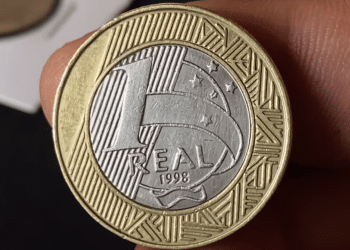 Close detalhado do reverso de uma moeda de 1 real de 1998, mostrando o ano e o valor, sendo segurada por dedos humanos para destacar o estado de conservação.