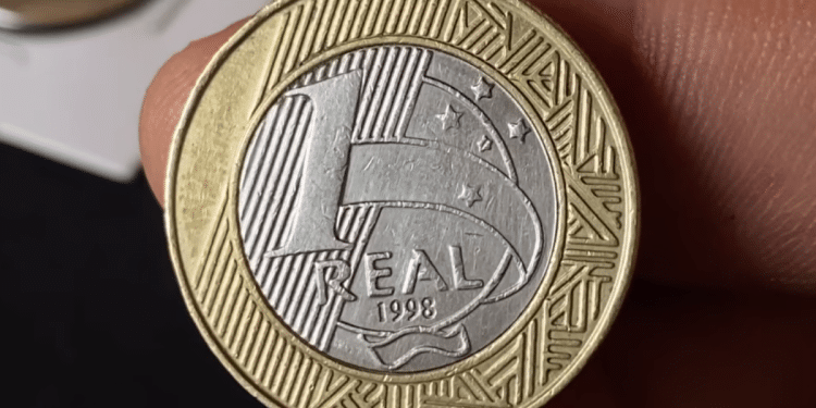 Close detalhado do reverso de uma moeda de 1 real de 1998, mostrando o ano e o valor, sendo segurada por dedos humanos para destacar o estado de conservação.