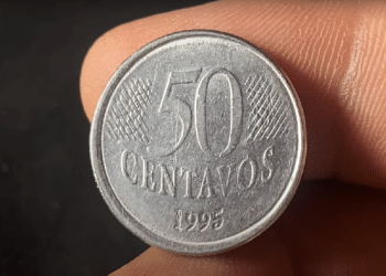 Close-up de uma mão segurando uma moeda de 50 centavos de 1995, em prata, contra um fundo escuro, focando nos detalhes do ano de cunhagem.