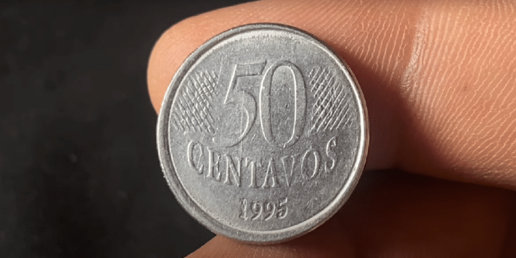 Close-up de uma mão segurando uma moeda de 50 centavos de 1995, em prata, contra um fundo escuro, focando nos detalhes do ano de cunhagem.
