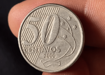 Close nítido de uma moeda de 50 centavos do Brasil, ano 2000, destacando o numeral 50 e as estrelas do Cruzeiro do Sul, segurada entre os dedos para inspeção de raridade.