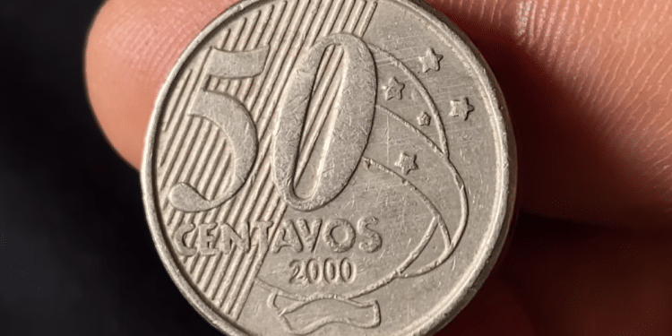 Close nítido de uma moeda de 50 centavos do Brasil, ano 2000, destacando o numeral 50 e as estrelas do Cruzeiro do Sul, segurada entre os dedos para inspeção de raridade.