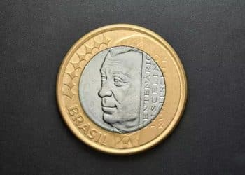 Close do anverso de uma moeda de 1 real comemorativa do Centenário de Juscelino Kubitschek, exibindo a efígie de JK no centro de prata com o anel dourado externo.