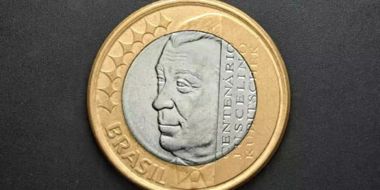 Close do anverso de uma moeda de 1 real comemorativa do Centenário de Juscelino Kubitschek, exibindo a efígie de JK no centro de prata com o anel dourado externo.