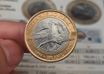Mão segurando uma moeda de 1 real comemorativa de 25 anos do Real, com o desenho do Beija-Flor, sobre um catálogo de preços de numismática.