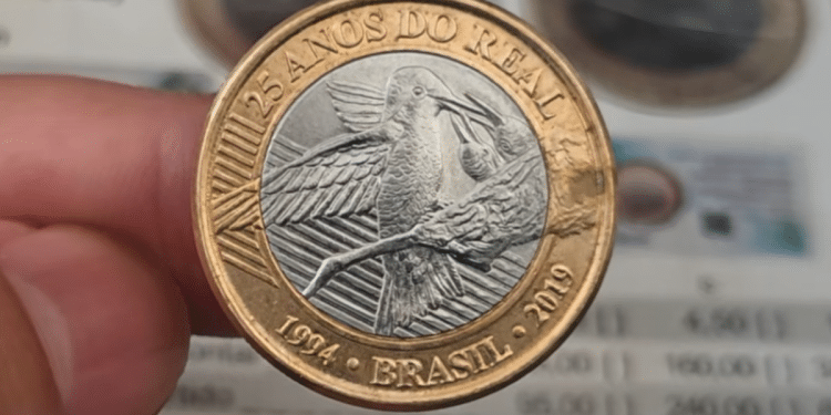 Mão segurando uma moeda de 1 real comemorativa de 25 anos do Real, com o desenho do Beija-Flor, sobre um catálogo de preços de numismática.