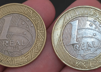Foto em primeiro plano de duas moedas brasileiras de 1 real sendo seguradas entre os dedos. A moeda da esquerda mostra o ano de 1998 e a da direita exibe o ano de 1999, ambas com o brilho metálico preservado.
