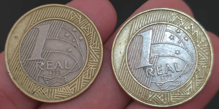 Foto em primeiro plano de duas moedas brasileiras de 1 real sendo seguradas entre os dedos. A moeda da esquerda mostra o ano de 1998 e a da direita exibe o ano de 1999, ambas com o brilho metálico preservado.