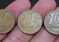 Close-up de três moedas de 10 centavos da segunda família do Real, das datas 1998, 1999 e 2000, sendo seguradas entre os dedos sobre um fundo escuro.