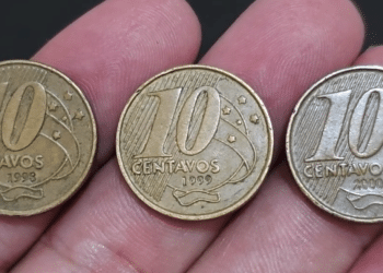 Close-up de três moedas de 10 centavos da segunda família do Real, das datas 1998, 1999 e 2000, sendo seguradas entre os dedos sobre um fundo escuro.