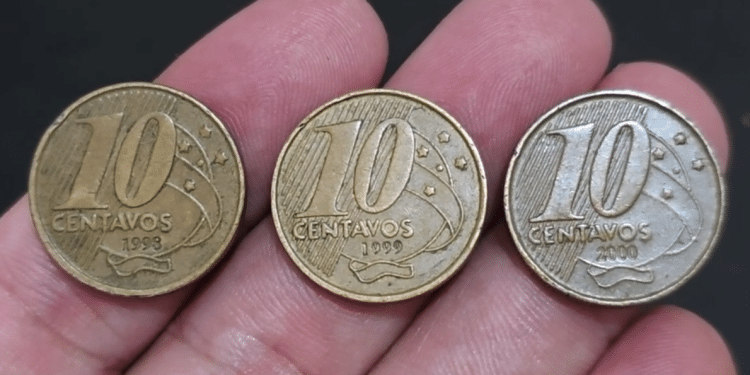 Close-up de três moedas de 10 centavos da segunda família do Real, das datas 1998, 1999 e 2000, sendo seguradas entre os dedos sobre um fundo escuro.