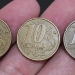 Close-up de três moedas de 10 centavos da segunda família do Real, das datas 1998, 1999 e 2000, sendo seguradas entre os dedos sobre um fundo escuro.