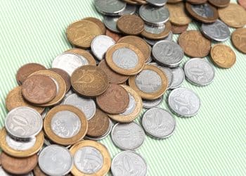 Close-up de moedas brasileiras de real em ângulo inclinado sobre um fundo verde listrado. A imagem foca nas moedas de 50 centavos, 25 centavos, 10 centavos e 1 real, mostrando detalhes claros da inscrição 'CENTAVOS' e 'REAL' e os designs de cunhagem. As moedas estão amontoadas e em perspectiva, com o foco diminuindo para as moedas mais distantes.