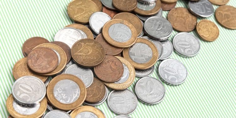 Close-up de moedas brasileiras de real em ângulo inclinado sobre um fundo verde listrado. A imagem foca nas moedas de 50 centavos, 25 centavos, 10 centavos e 1 real, mostrando detalhes claros da inscrição 'CENTAVOS' e 'REAL' e os designs de cunhagem. As moedas estão amontoadas e em perspectiva, com o foco diminuindo para as moedas mais distantes.