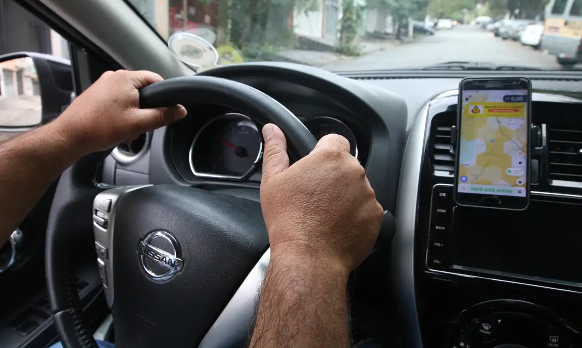 Mãos de motorista segurando o volante de um carro Nissan em movimento, com smartphone fixado no painel exibindo aplicativo de navegação com mapa e status online, representando a ocupação de motorista de transporte por aplicativos reconhecida oficialmente na Classificação Brasileira de Ocupações em 2026