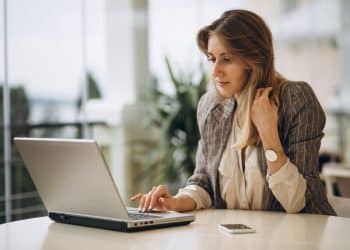 Mulher loira, de blazer listrado e relógio dourado, sentada à mesa e concentrada no uso de um notebook prateado.