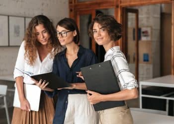 Três jovens mulheres profissionais em um escritório moderno; uma à direita segura uma prancheta e olha para a câmera sorrindo, enquanto as outras duas ao centro e à esquerda consultam juntas um notebook e uma pasta.
